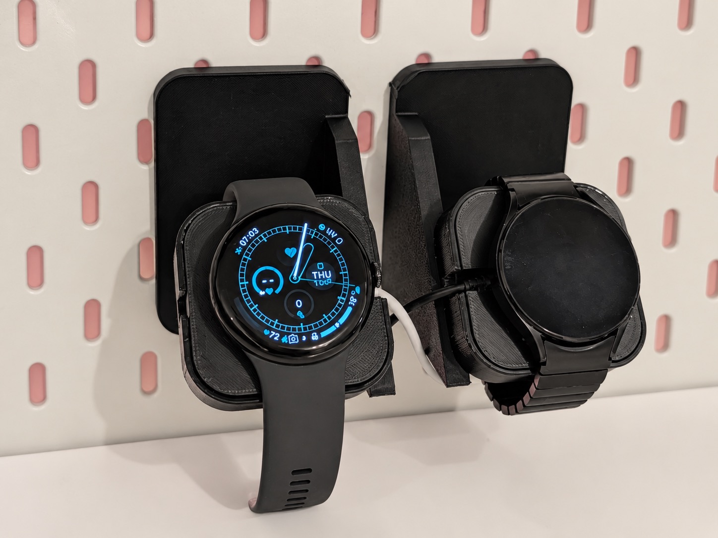 JDC Duo watch docks mounted on a pegboard, holding a Pixel Watch and Galaxy Watch